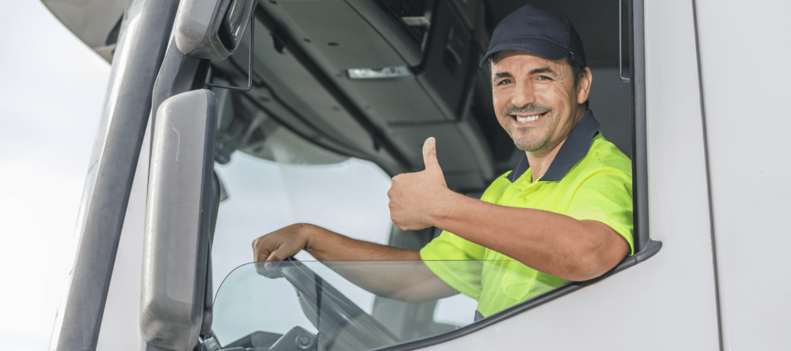 an hgv driver in high-vis is sitting in a white vehicle and making a thumbs up sign.