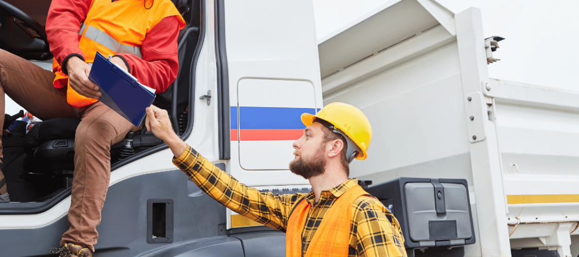 two hgv drivers are chatting over a clipboard. they both wear high vis and hard hats. one is standing on the ground and the other is in the lorry cab.