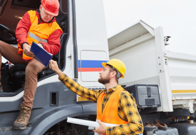 two hgv drivers are chatting over a clipboard. they both wear high vis and hard hats. one is standing on the ground and the other is in the lorry cab.